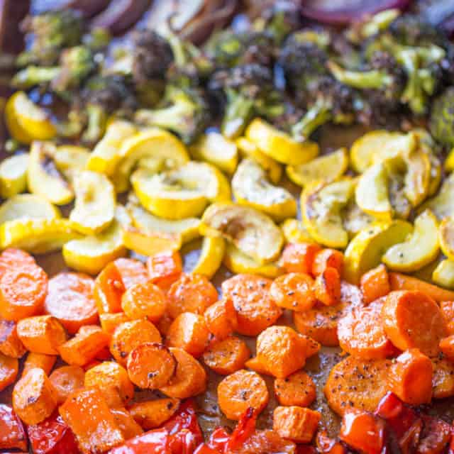 Rainbow roasted vegetables lined up on a baking sheet, including carrots, bell peppers, zucchini, and red onions.